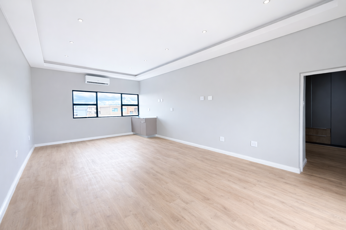 A bright, spacious, and modern living area featuring light wood-look flooring, recessed ceiling lighting, a wall-mounted air conditioning unit, and a small built-in kitchenette.