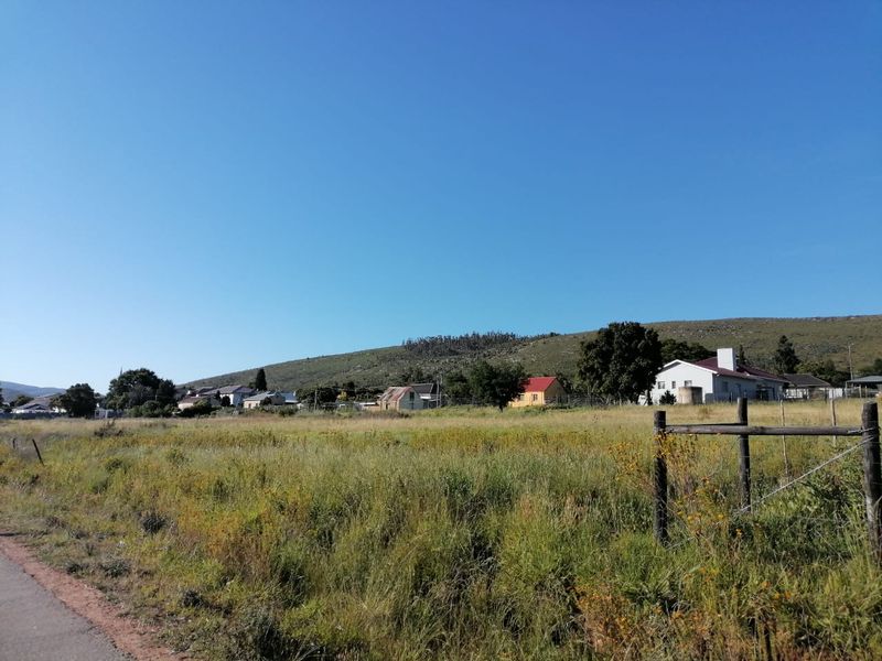 A view of a vacant residential plot featuring natural grass cover, enclosed by wire fencing with wooden posts, set against a hillside backdrop.