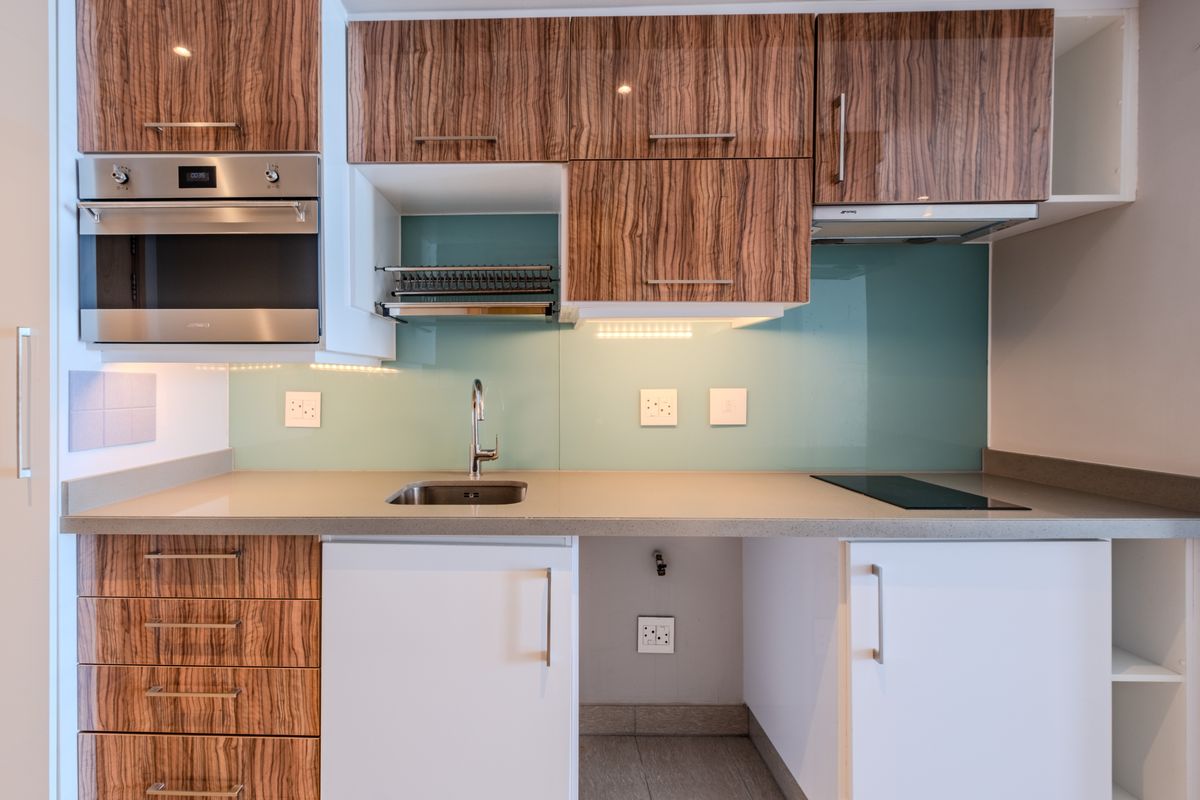 Modern kitchen featuring wood-grain cabinetry, stone countertops, a glass splashback, built-in oven, and integrated hob with under-cabinet lighting.