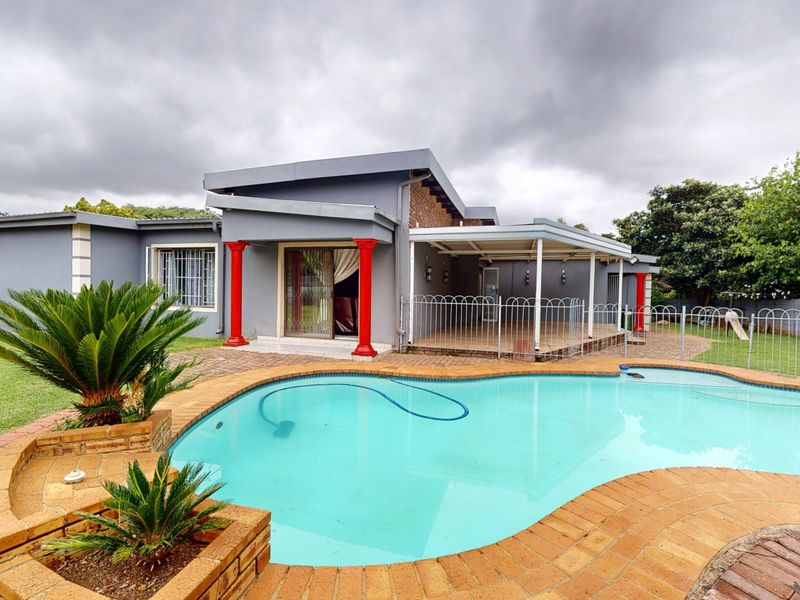 Rear exterior view featuring a swimming pool with brick coping, a covered patio area, and decorative pillars set against a landscaped garden.