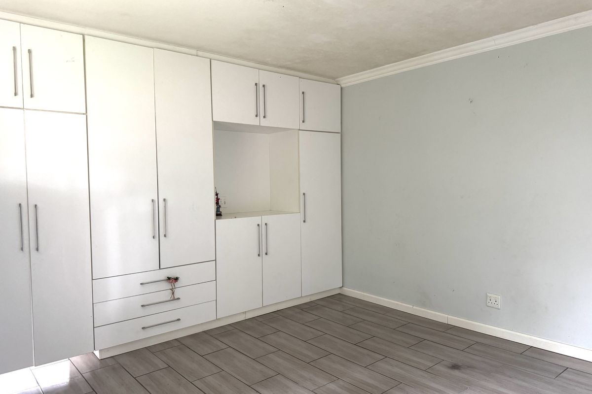 A bright bedroom featuring extensive white built-in cupboards, modern wood-look tiled flooring, and neutral wall finishes.