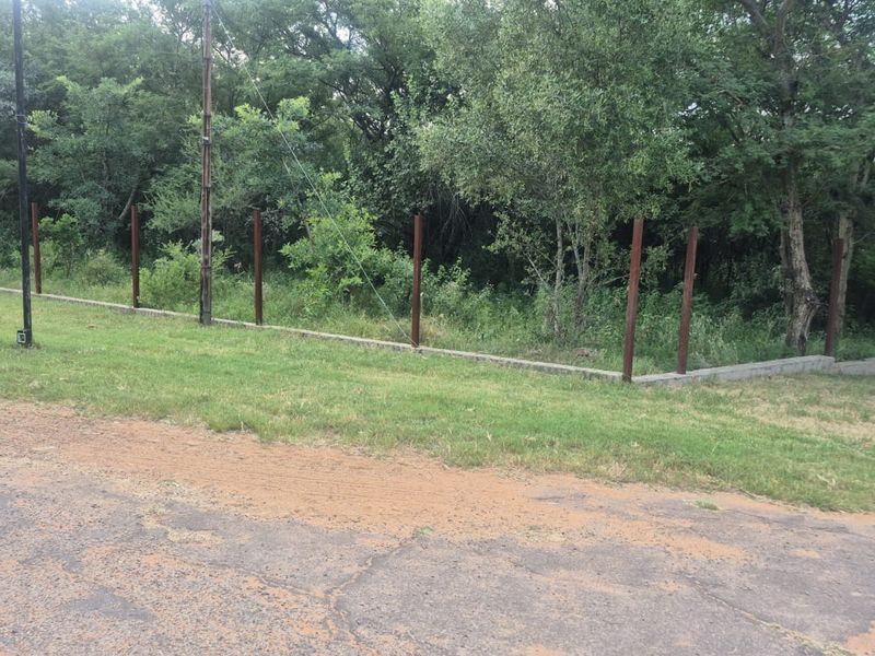 A view of a vacant residential plot featuring a concrete retaining wall and metal fence posts bordering a wooded area.