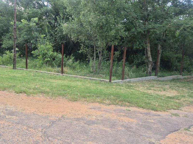 A view of the vacant residential plot featuring a concrete perimeter wall and metal fence posts bordering a wooded area.