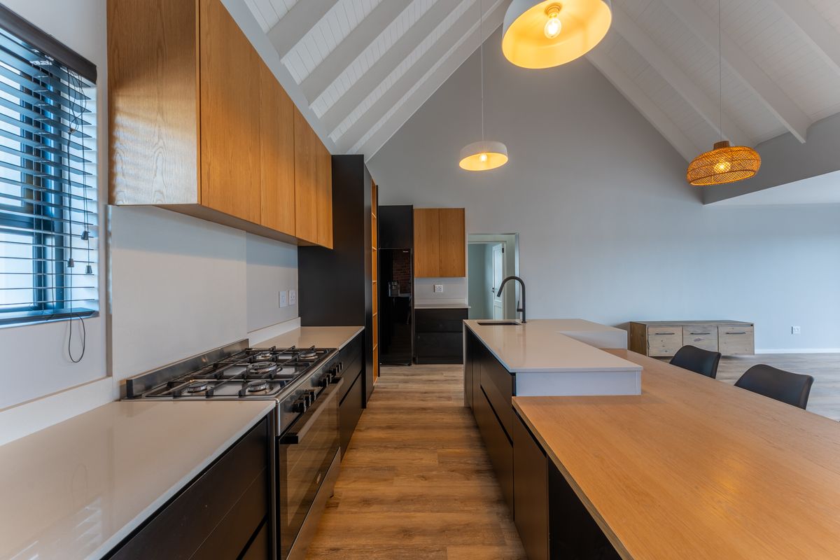 Modern kitchen featuring timber cabinetry, stone countertops, a gas hob, and a central island under a high vaulted ceiling with pendant lighting.