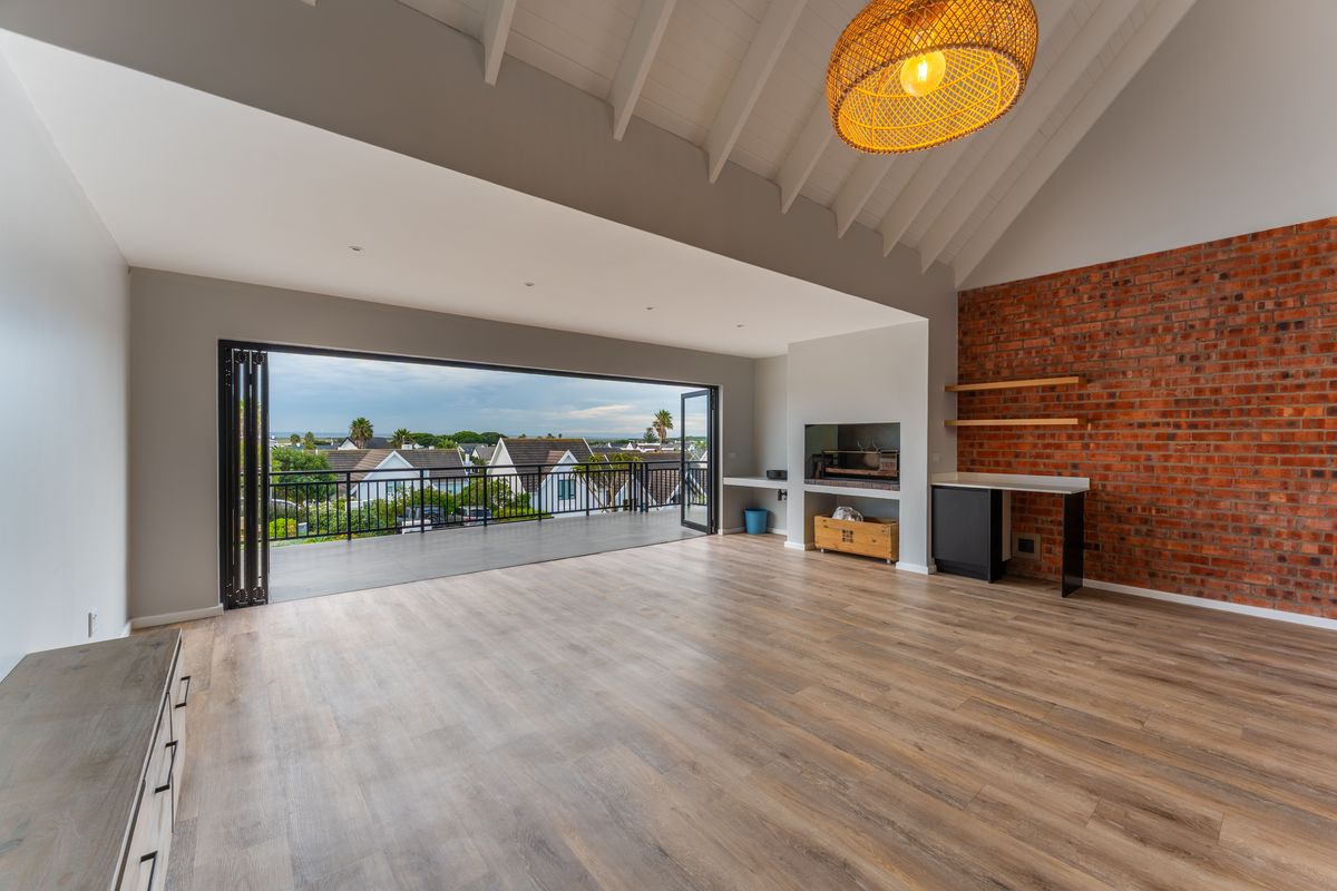 Spacious open-plan living area featuring a vaulted ceiling, exposed brick wall, built-in braai, and large bi-fold doors opening onto a balcony with scenic views.