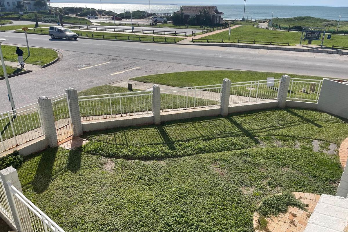 Elevated view of a private rear garden featuring a lawn, paved patio area, and secure boundary wall with metal fencing overlooking the coastal road.
