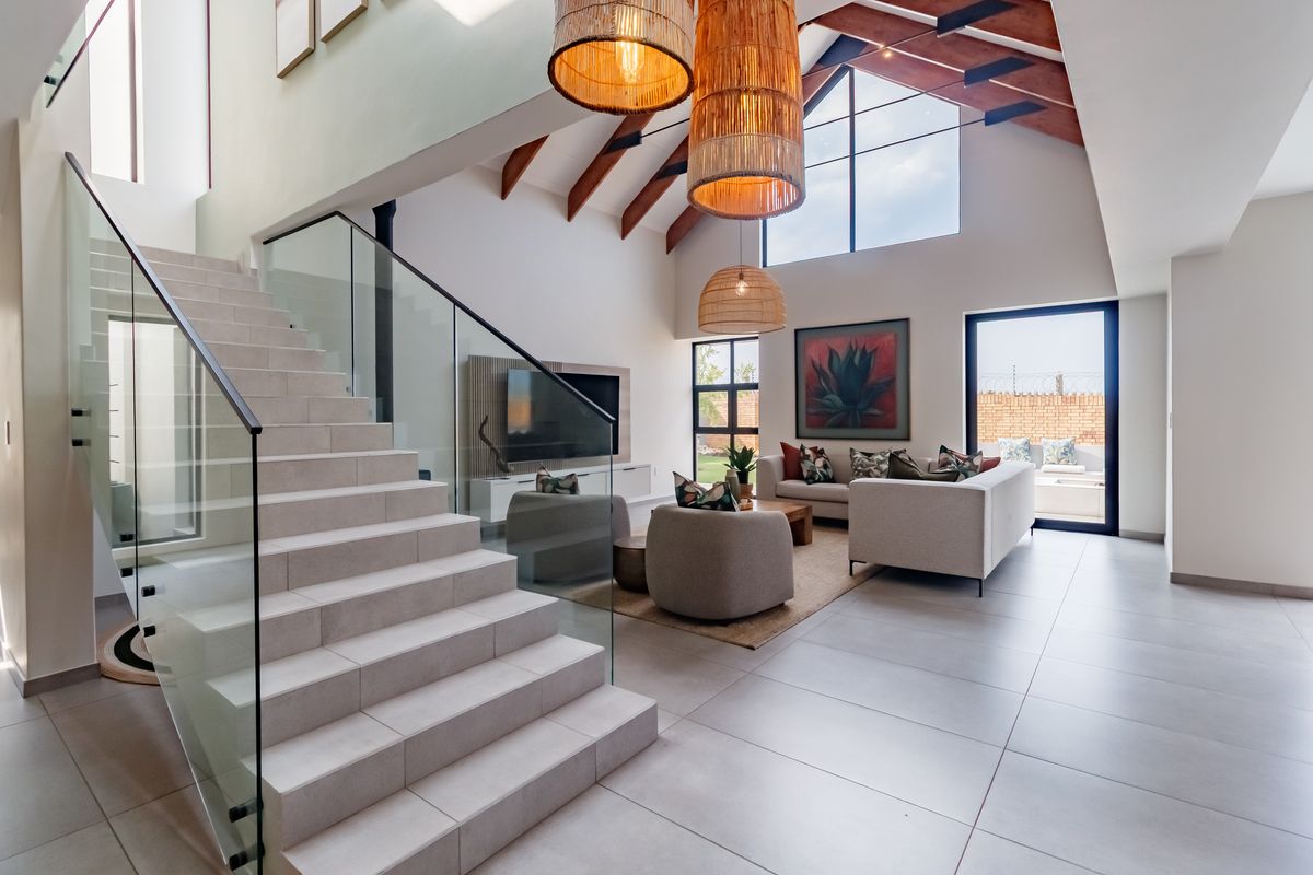 Spacious double-volume living area featuring exposed timber rafters, large windows, tiled flooring, and a modern staircase with a glass balustrade.