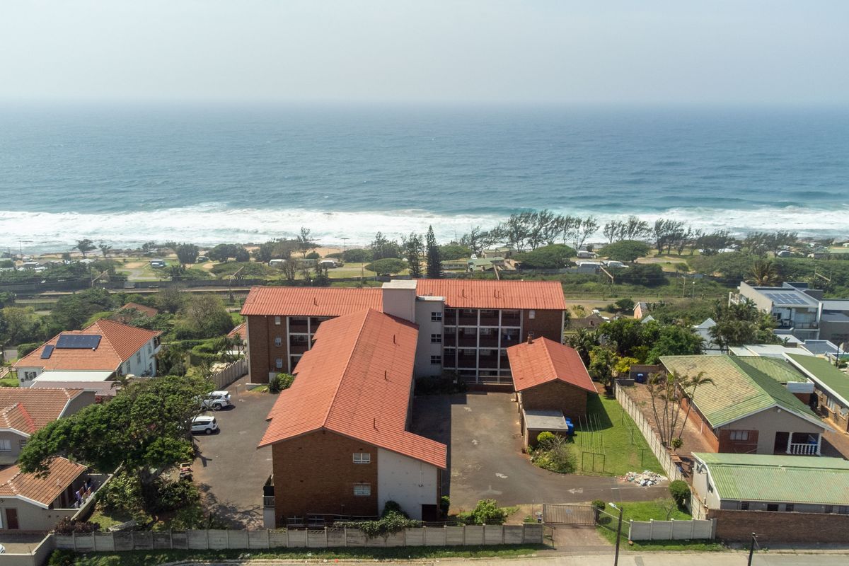 Aerial view of a multi-story residential apartment complex featuring a tiled roof, paved parking area, and proximity to the coastline.