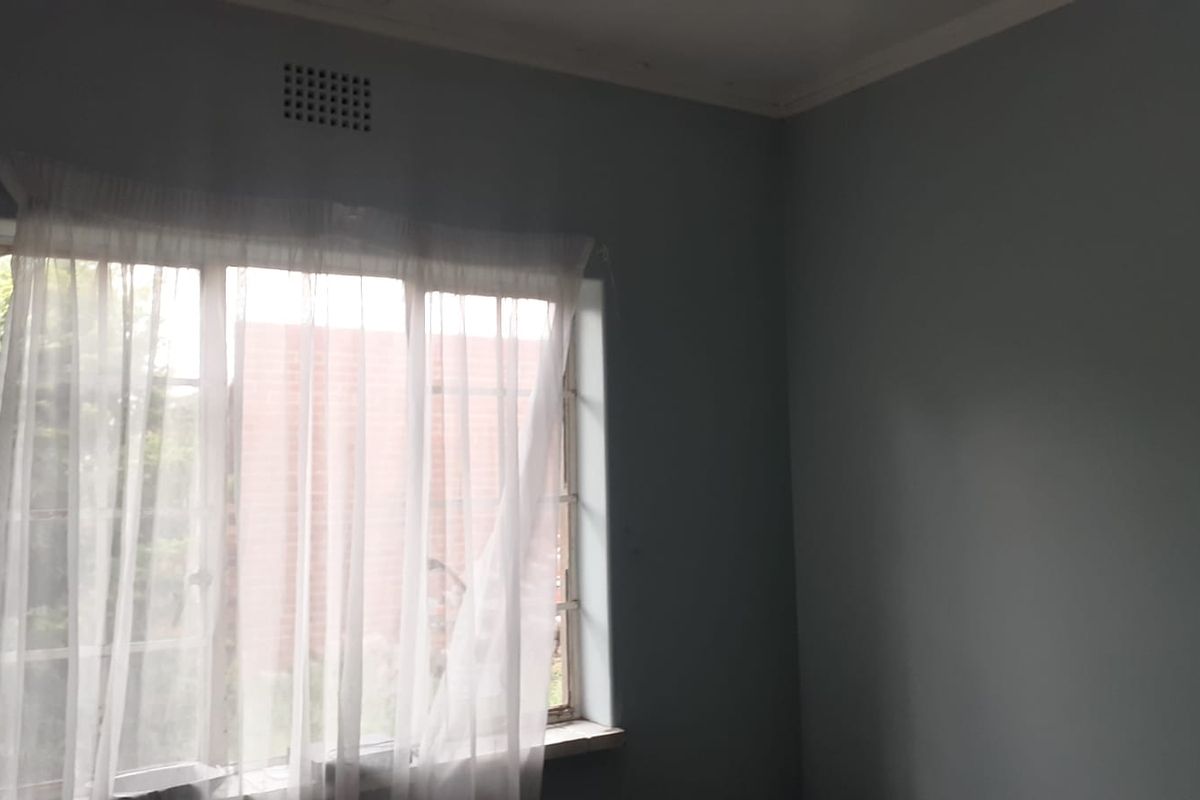 A simple bedroom featuring a large window with natural light, neutral wall tones, and a built-in ventilation vent.