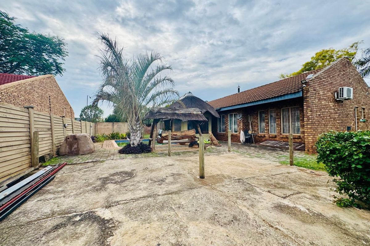 Rear exterior view of a brick house featuring a paved patio area, a thatched lapa, and a swimming pool in the background.