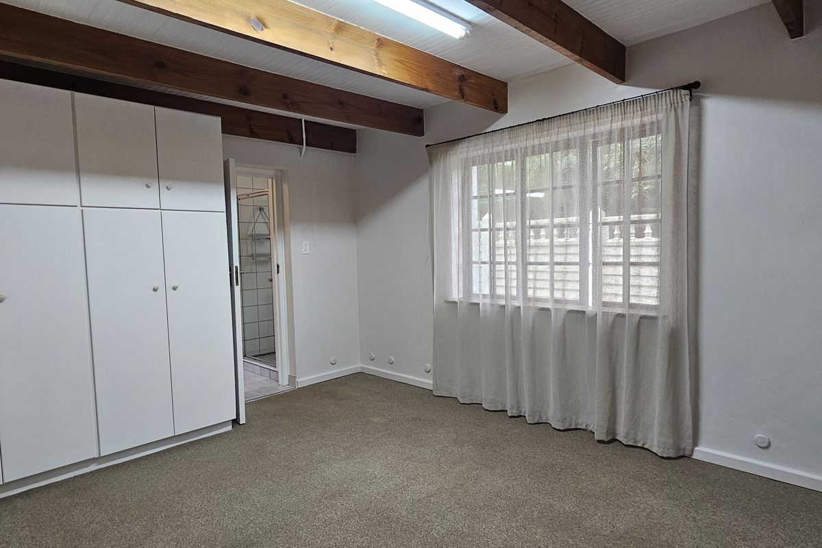 A spacious bedroom featuring exposed timber ceiling beams, neutral carpeting, and extensive built-in cupboard storage, with an adjoining en-suite bathroom.