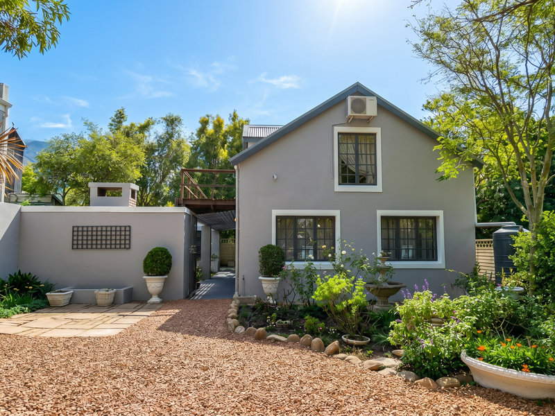 A well-maintained rear exterior view featuring a stone patio, gravel driveway, landscaped garden, and a timber balcony attached to the main structure.