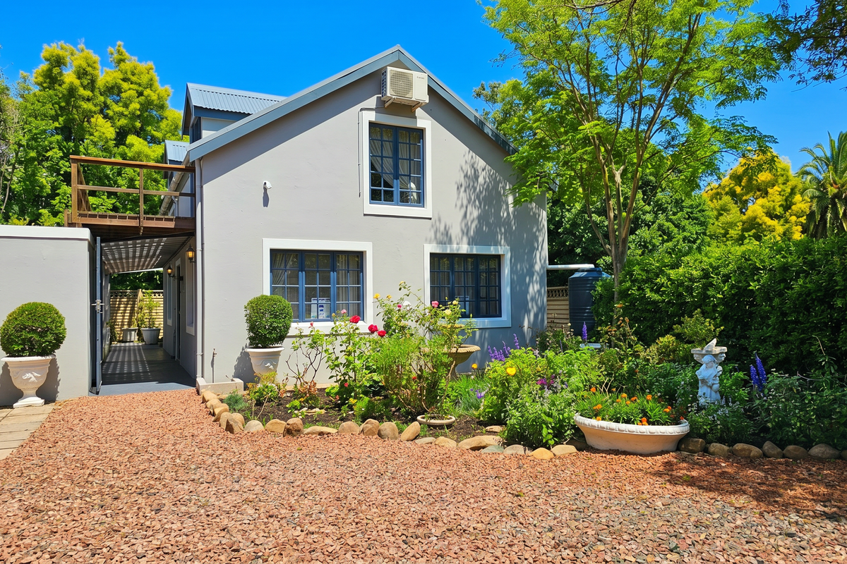 A well-maintained rear exterior view of the house featuring a timber balcony, gravel driveway, and established garden beds.