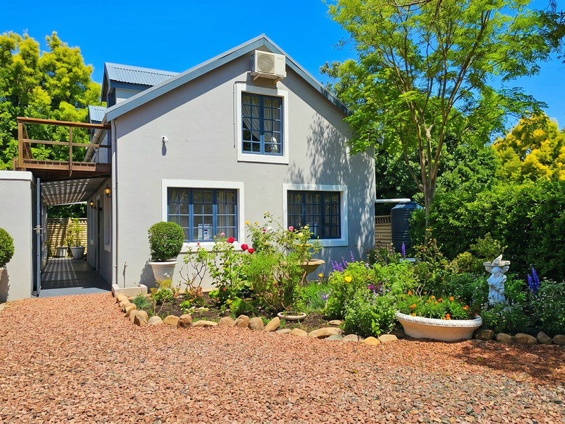 A well-maintained rear exterior view of the house featuring a timber balcony, gravel driveway, and established garden beds.