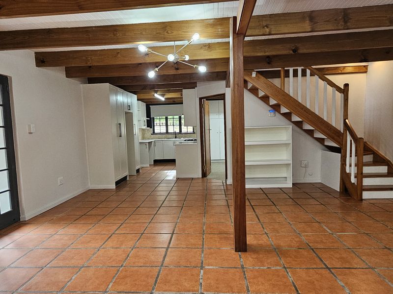Spacious open-plan living area featuring exposed timber beams, terracotta tile flooring, and a central staircase, seamlessly connecting to the kitchen space.