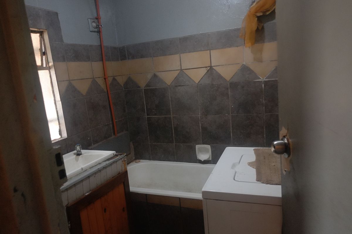 A dated bathroom featuring a built-in bathtub, vanity basin, and exposed plumbing, requiring cosmetic updates and modernization of finishes.
