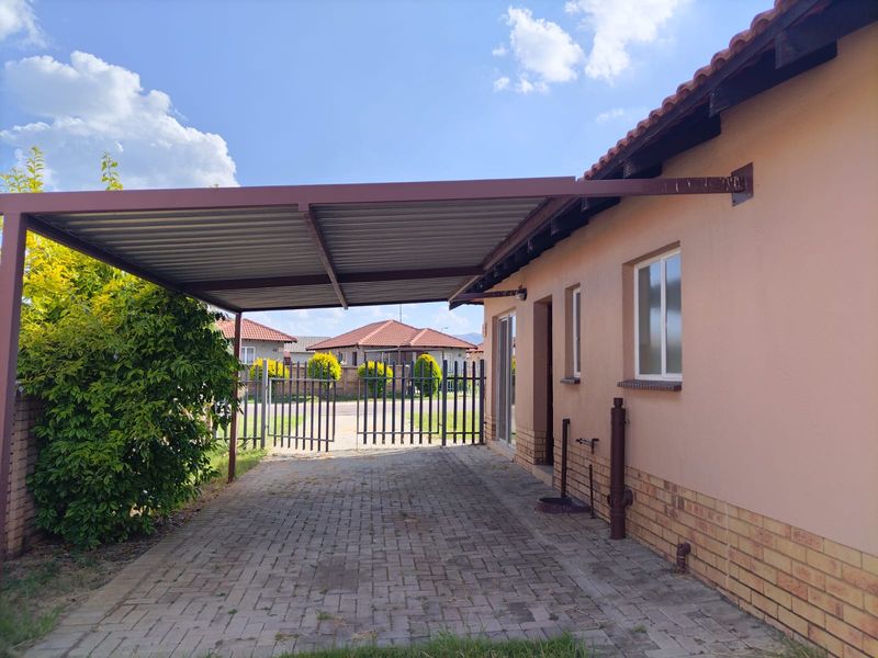 A paved driveway featuring a covered carport structure, brick plinth detailing, and a metal security gate providing access to the property.