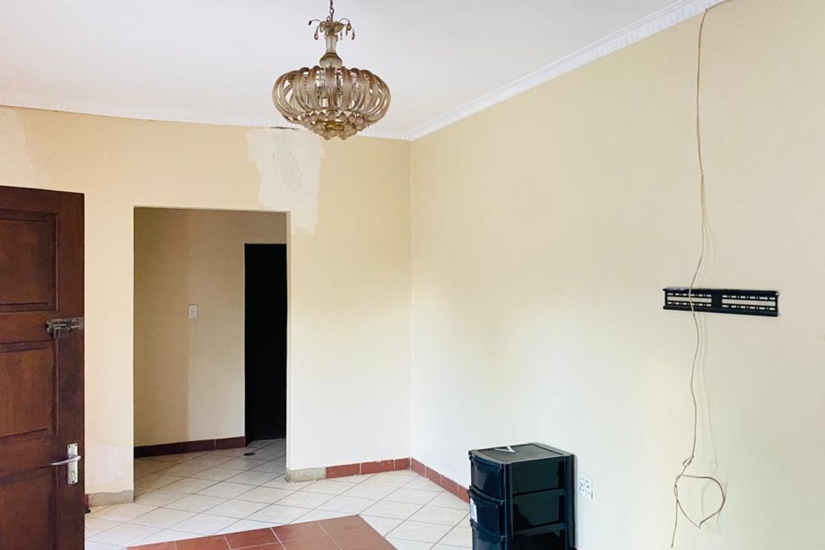 An empty living room featuring tiled flooring, decorative coving, and a central chandelier, currently requiring cosmetic wall repairs and maintenance.
