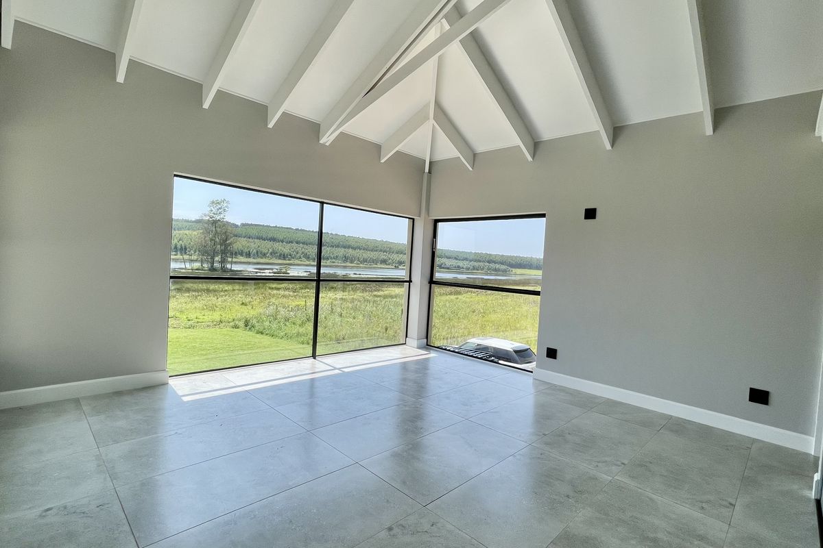 A bright, modern bedroom featuring high vaulted ceilings with exposed beams, large floor-to-ceiling windows with scenic views, and sleek tiled flooring.