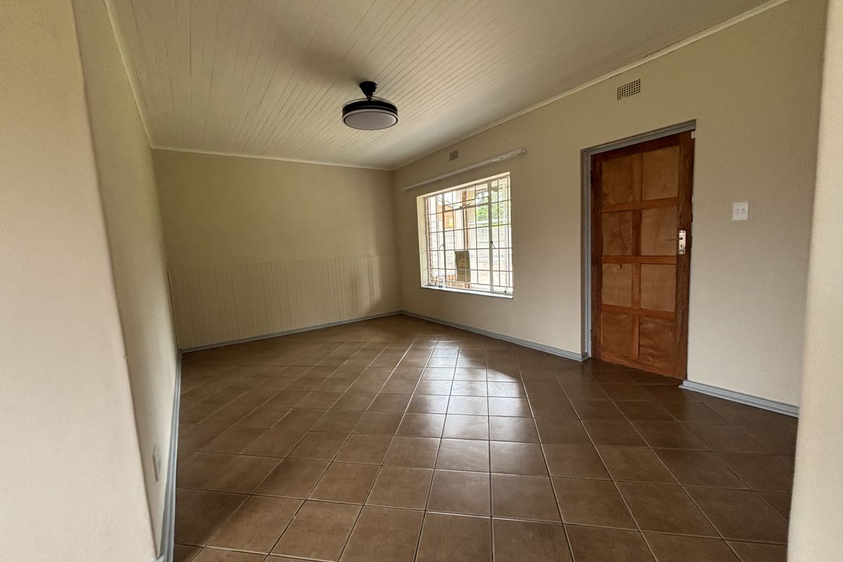 A spacious, empty living room featuring tiled flooring, a wooden door, and a window with security bars.