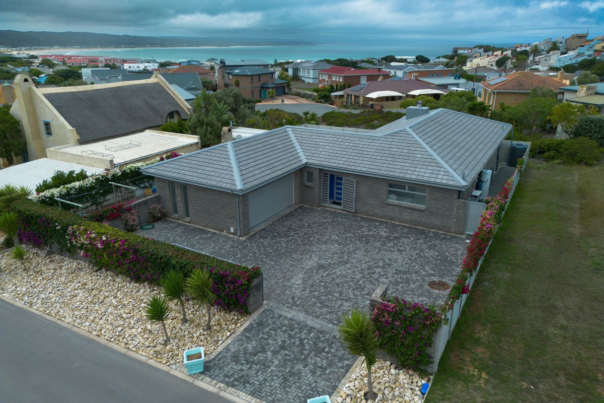 Aerial view of a modern brick house with a tiled roof, paved driveway, and landscaped boundary, situated in a coastal residential neighborhood.