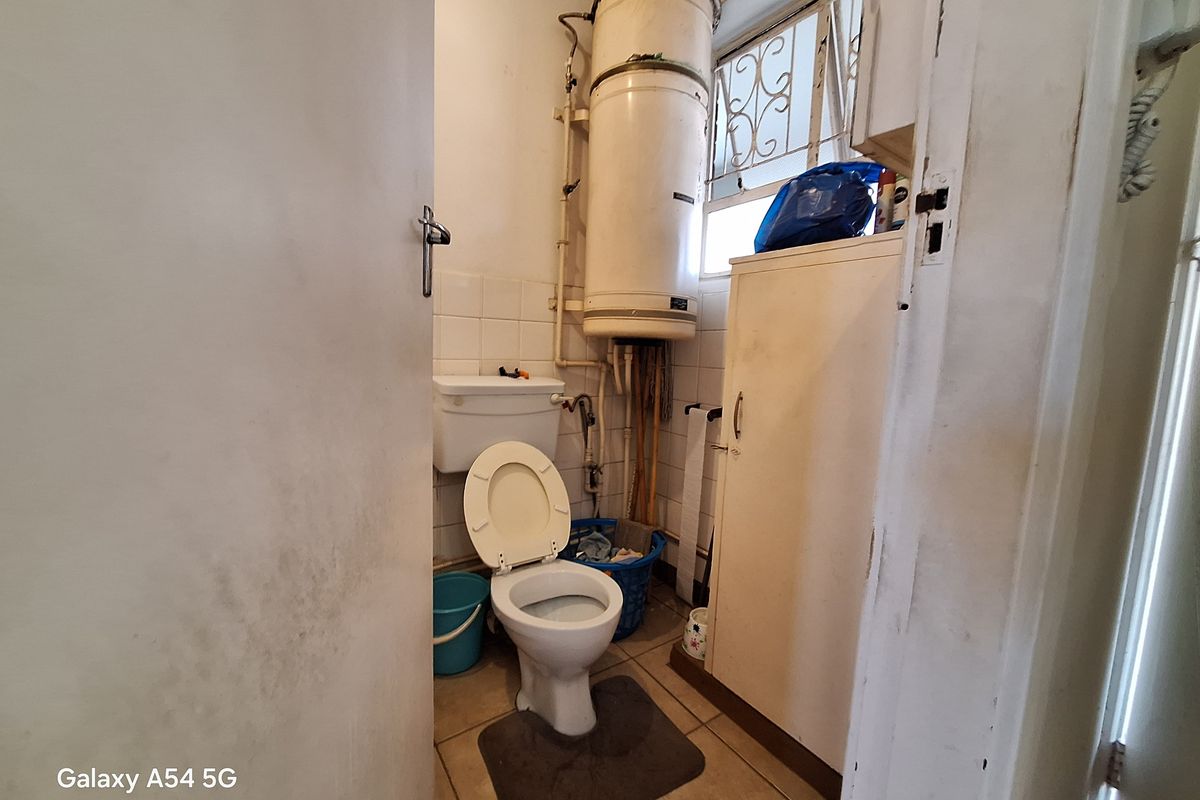 A compact bathroom featuring a toilet, wall-mounted geyser, and tiled flooring, requiring cosmetic updates and maintenance.