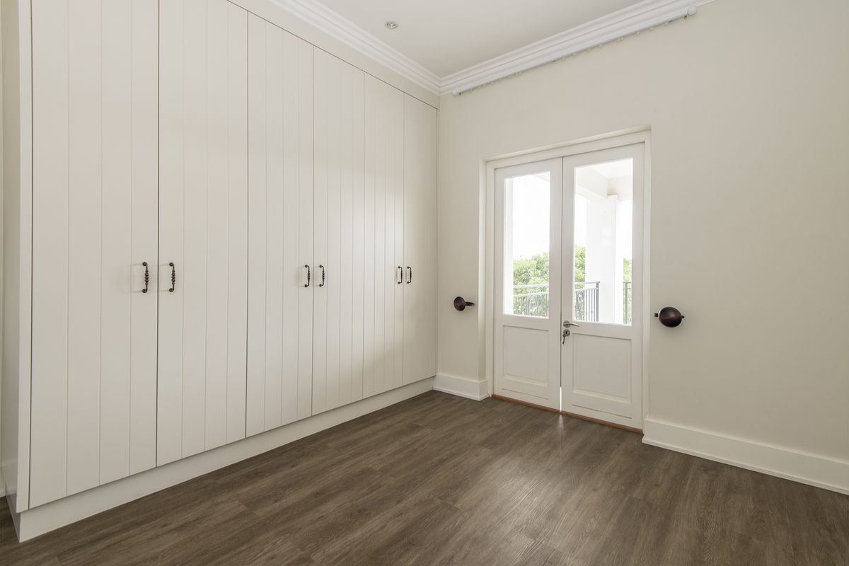 A bright bedroom featuring extensive floor-to-ceiling built-in cabinetry, wood-look flooring, crown molding, and double glass doors leading to an outdoor balcony.