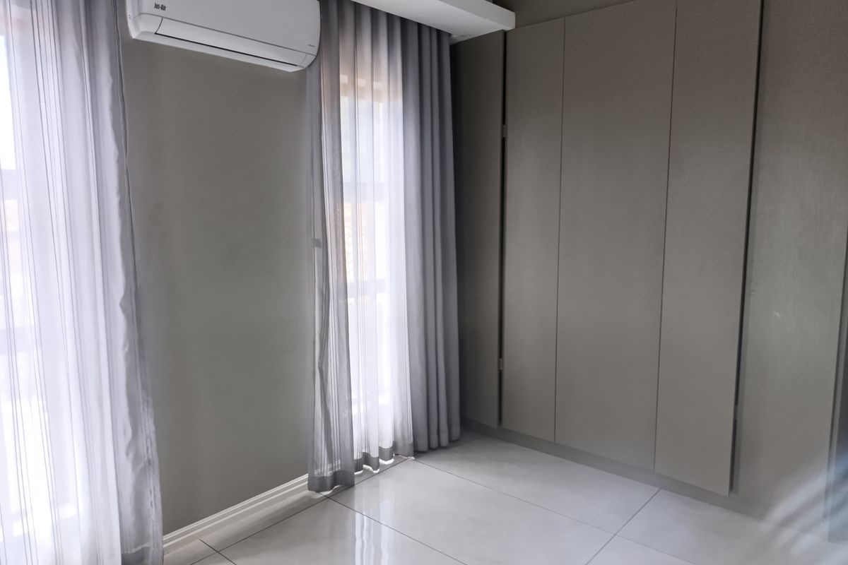 A clean, modern bedroom featuring neutral grey walls, large format floor tiles, built-in cupboards, and a wall-mounted air conditioning unit.