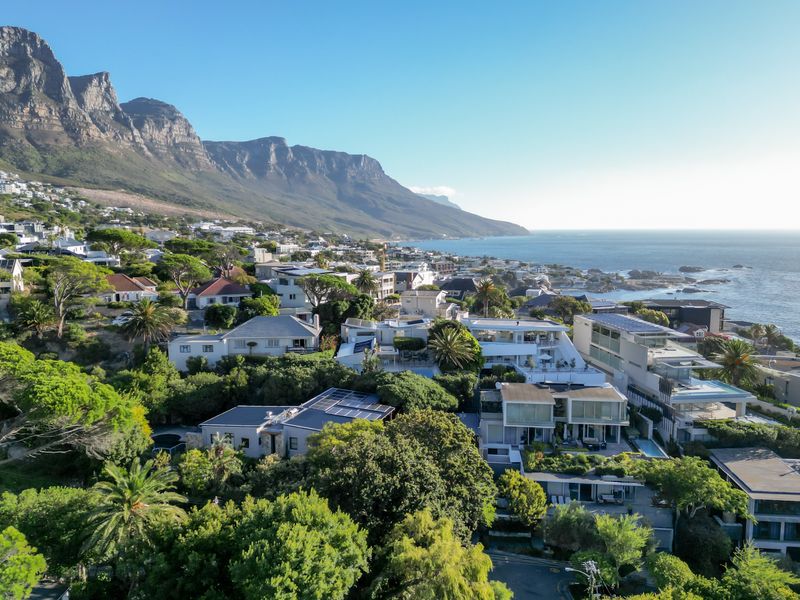 An elevated aerial view showcasing the coastal residential neighborhood, featuring lush greenery, modern homes with solar installations, and a stunning mountain and ocean backdrop.