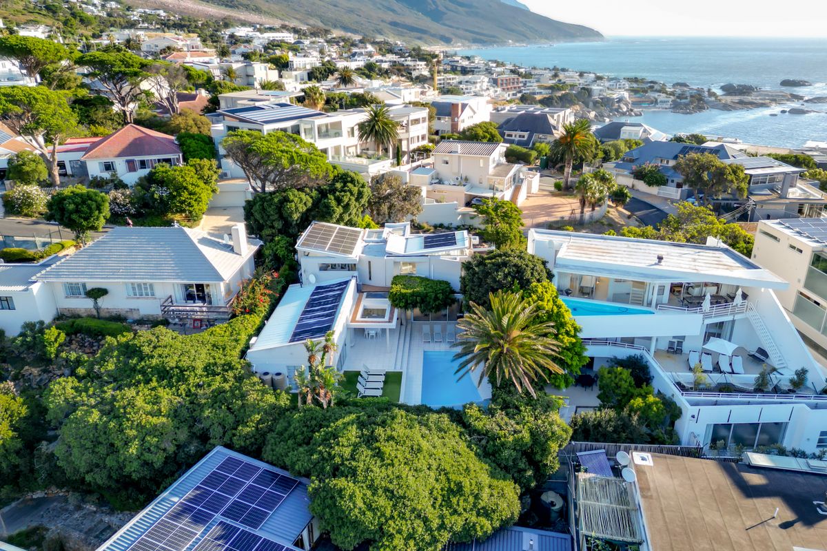 Aerial view of a coastal residential property featuring multiple roof levels, solar panels, and a private swimming pool set within a landscaped garden.