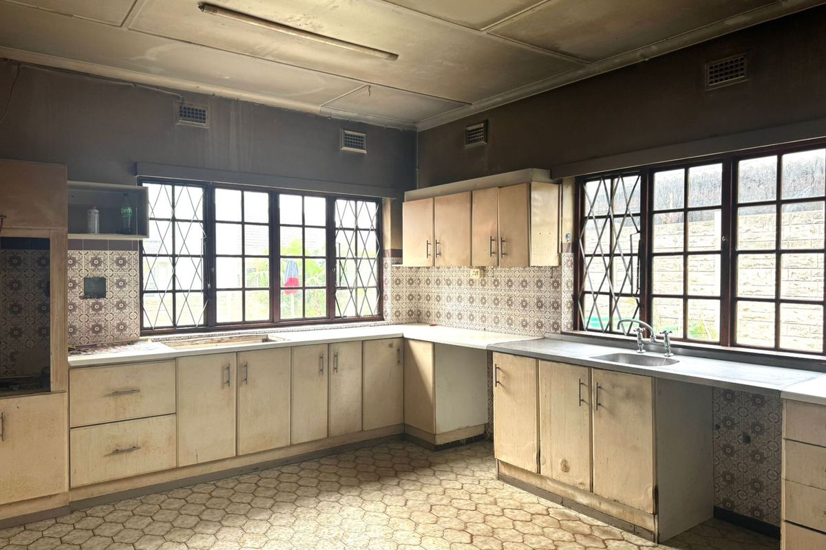 A dated, empty kitchen featuring original laminate cabinetry, tiled splashbacks, and large windows with security bars, offering significant potential for a modern renovation.