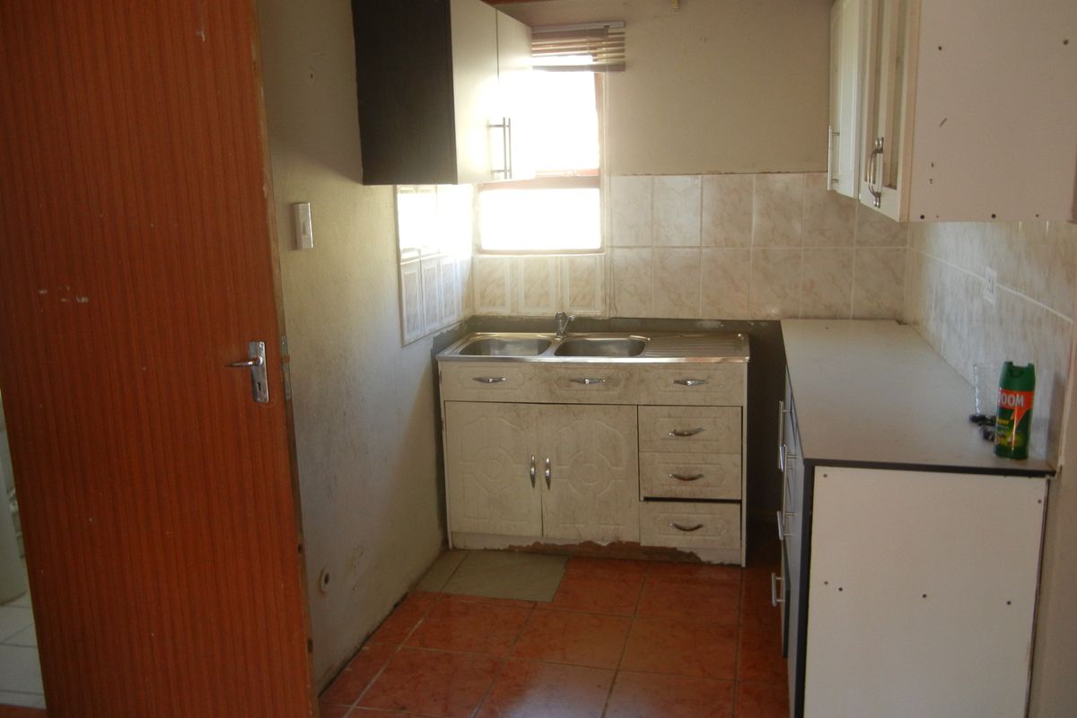 A small, dated kitchen featuring built-in cabinetry, a double sink, tiled splashbacks, and tiled flooring, requiring cosmetic updates and repairs.