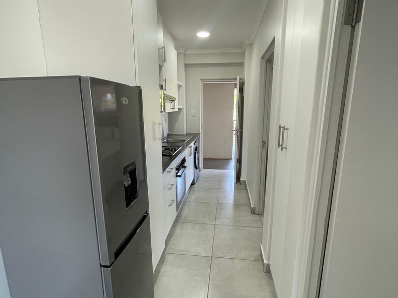 A galley-style kitchen featuring modern white cabinetry, tiled flooring, and integrated appliances, leading through to an adjacent room.