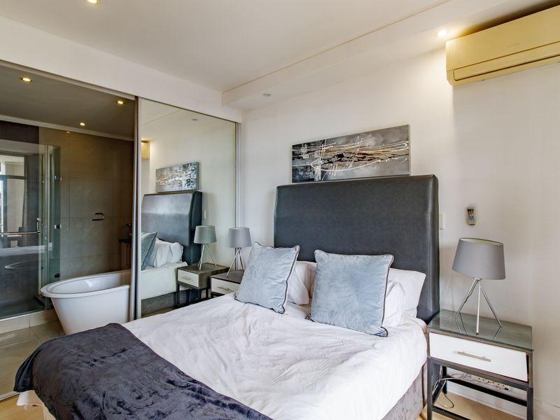 Modern bedroom featuring mirrored sliding wardrobes, recessed lighting, split-system air conditioning, and an open-plan en-suite bathroom with a freestanding bathtub.