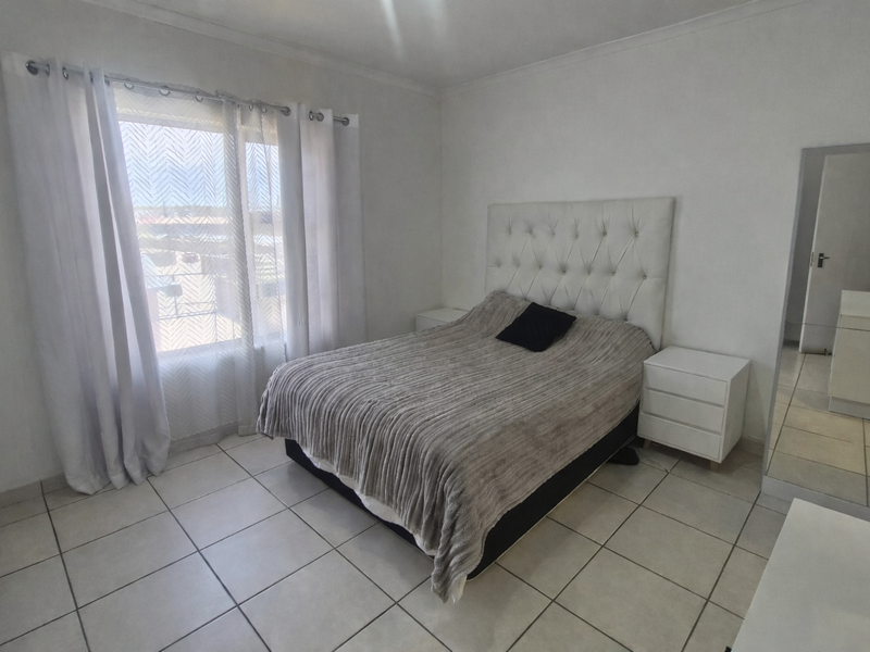 A bright bedroom featuring neutral tiled flooring, a built-in cupboard with a mirrored door, and large windows allowing for natural light.