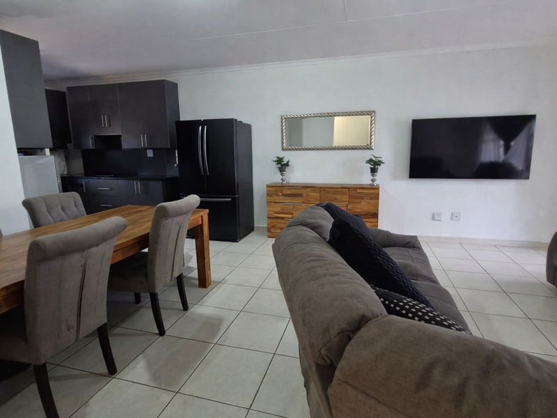 An open-plan living and dining area featuring tiled flooring, a modern kitchen with dark cabinetry, and a wall-mounted television.