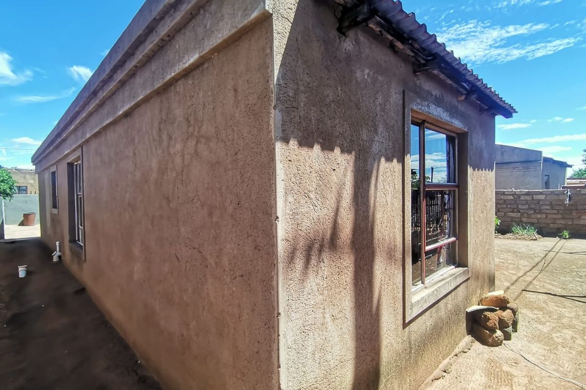 Rear exterior view of a single-story residential structure featuring plastered masonry walls, corrugated metal roofing, and steel-framed windows.