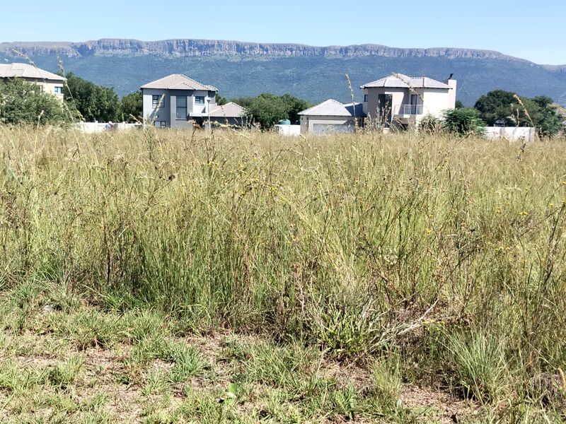 A vacant residential plot featuring tall grass with scenic mountain views and surrounding established residential properties.