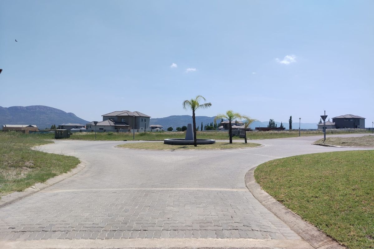 A view of a paved roundabout and landscaped central island within a residential estate, featuring surrounding vacant land and distant mountain views.