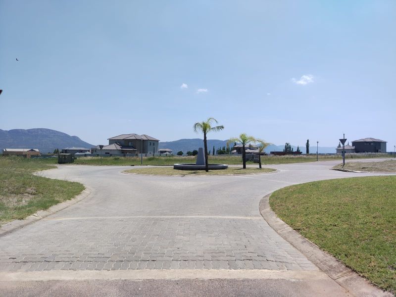 A view of a paved roundabout and landscaped central island within a residential estate, featuring surrounding vacant land and distant mountain views.