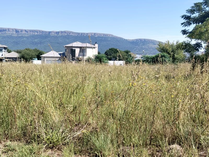 A view of a vacant residential plot featuring tall grass with surrounding neighborhood homes and mountain views in the background.