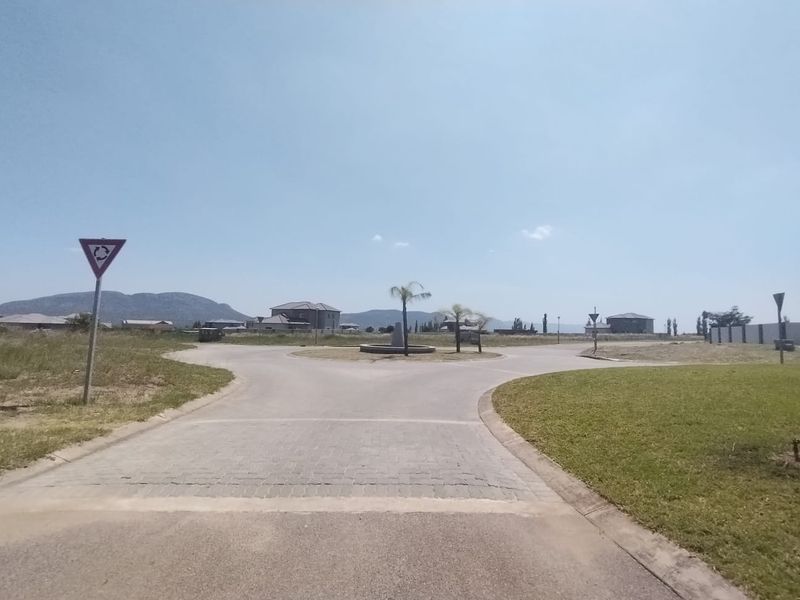 A view of a paved roundabout and surrounding residential land within a developed estate, featuring manicured lawns and distant mountain views.