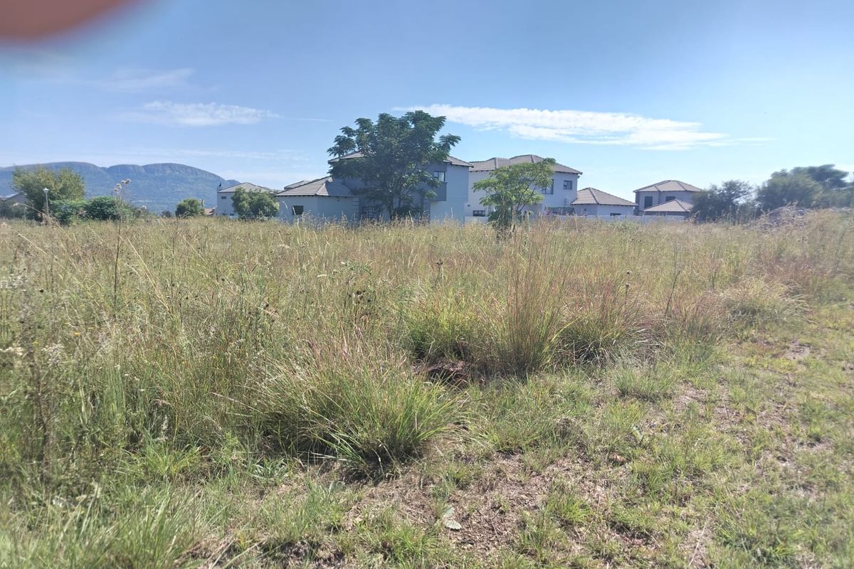 A view of a vacant residential plot featuring natural grass coverage with surrounding residential development and mountain views in the distance.