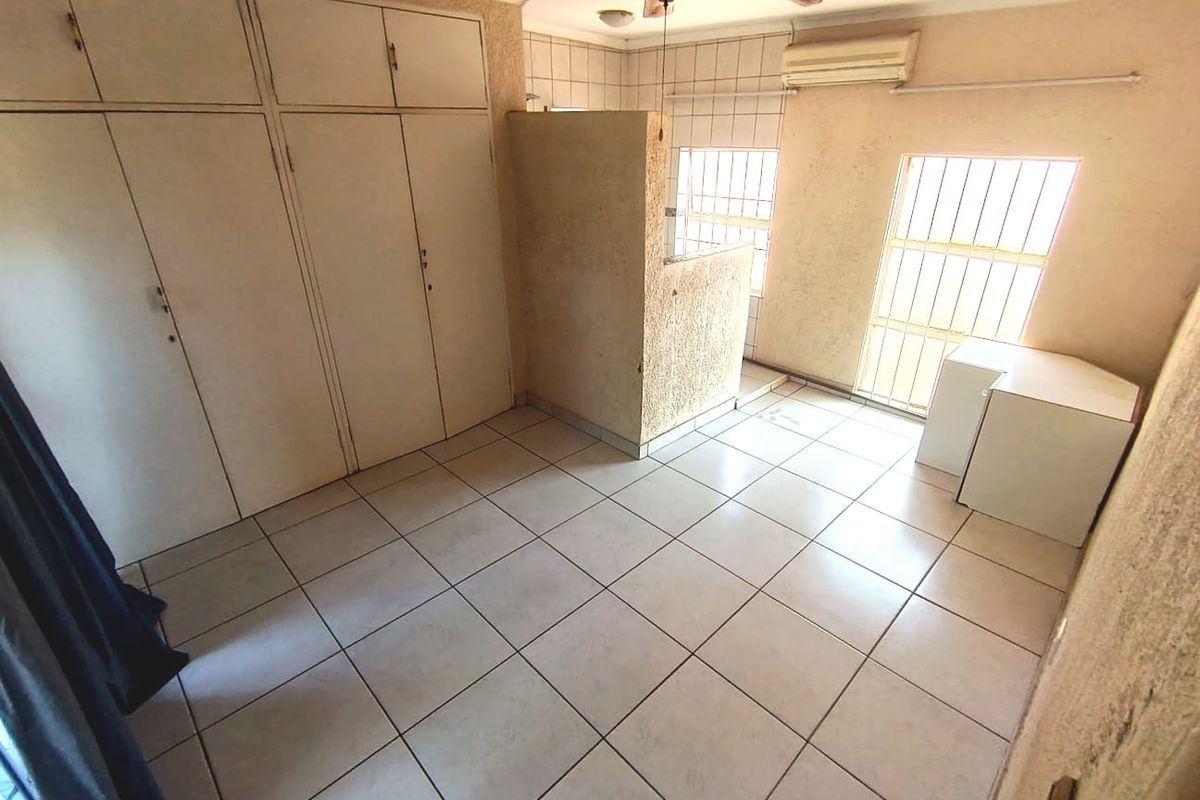 An empty bedroom featuring tiled flooring, built-in cupboards, a ceiling fan, and wall-mounted air conditioning, with security bars installed on the windows.