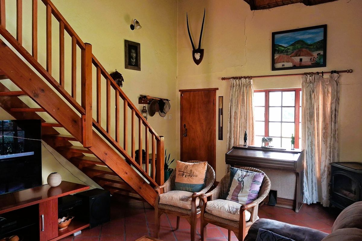 A rustic living room featuring a timber staircase, terracotta flooring, and a wood-burning stove under a traditional thatched ceiling.