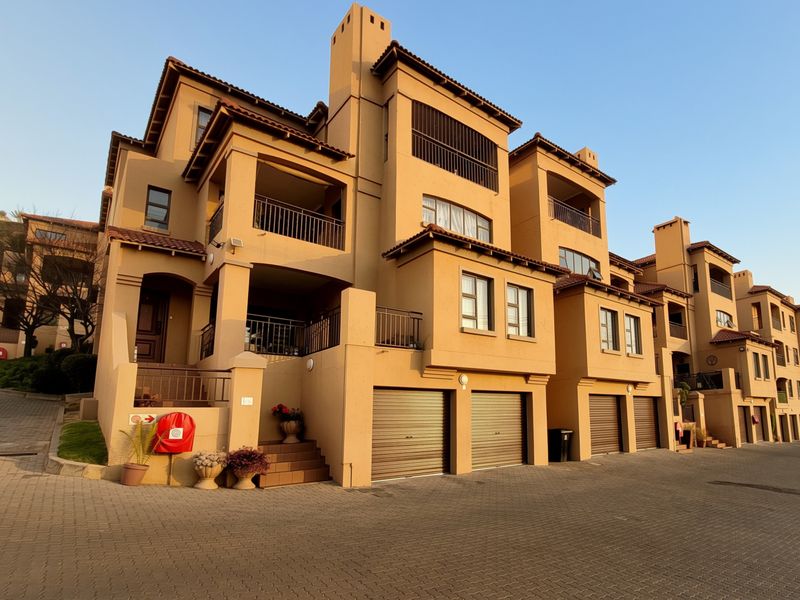 A multi-story apartment complex featuring a row of integrated garages, private balconies, and a paved driveway area under a clear sky.