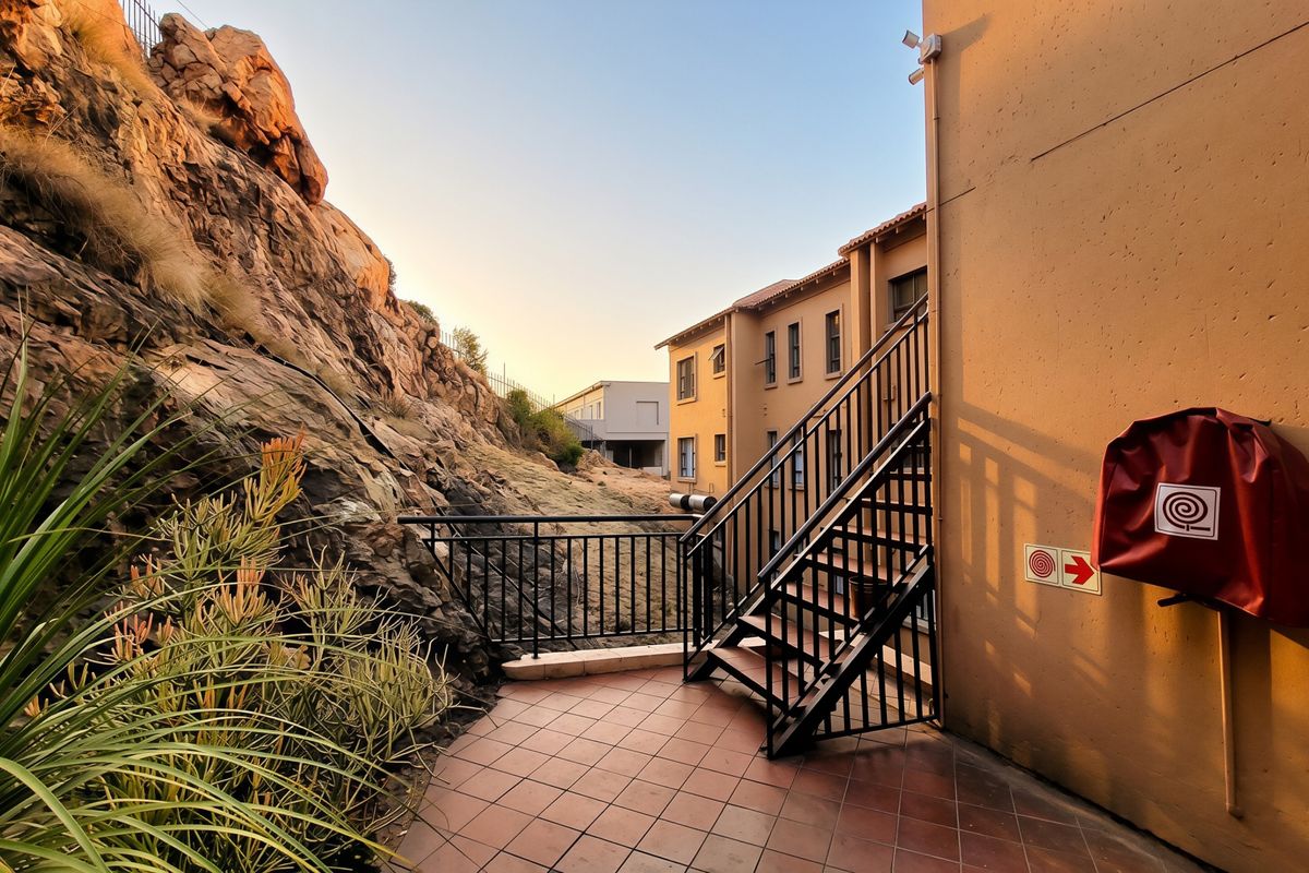 An exterior rear patio area featuring tiled flooring, a metal staircase, and a fire hose reel, set against a natural rock face.