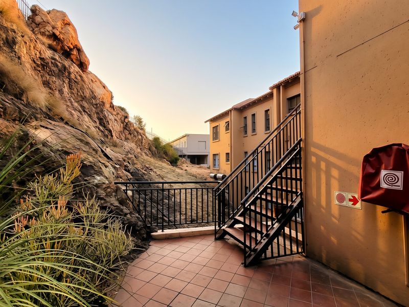 An exterior rear patio area featuring tiled flooring, a metal staircase, and a fire hose reel, set against a natural rock face.