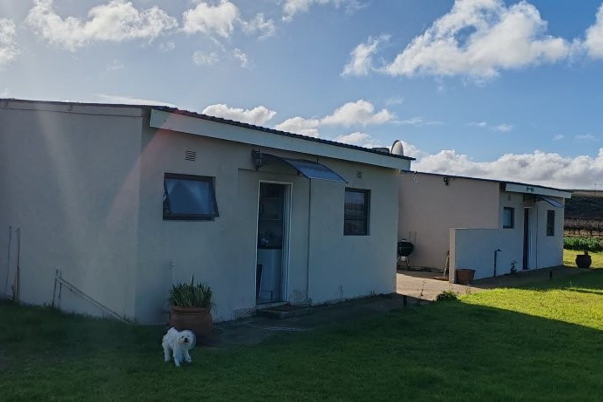 A rear view of a single-story farm dwelling featuring a chimney, paved patio area, and surrounding lawn with rural landscape views.