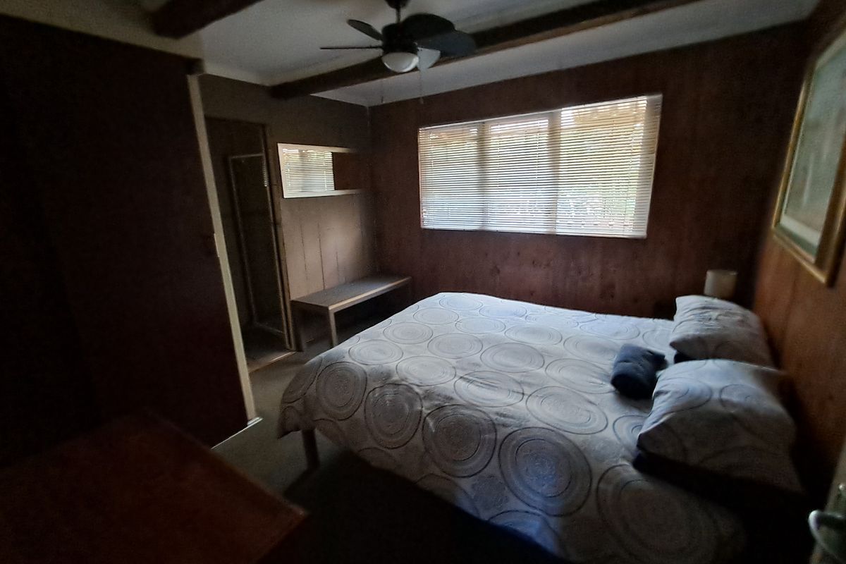 A bedroom featuring dark wood paneling, exposed ceiling beams, and a ceiling fan, offering a rustic aesthetic in need of modernization.